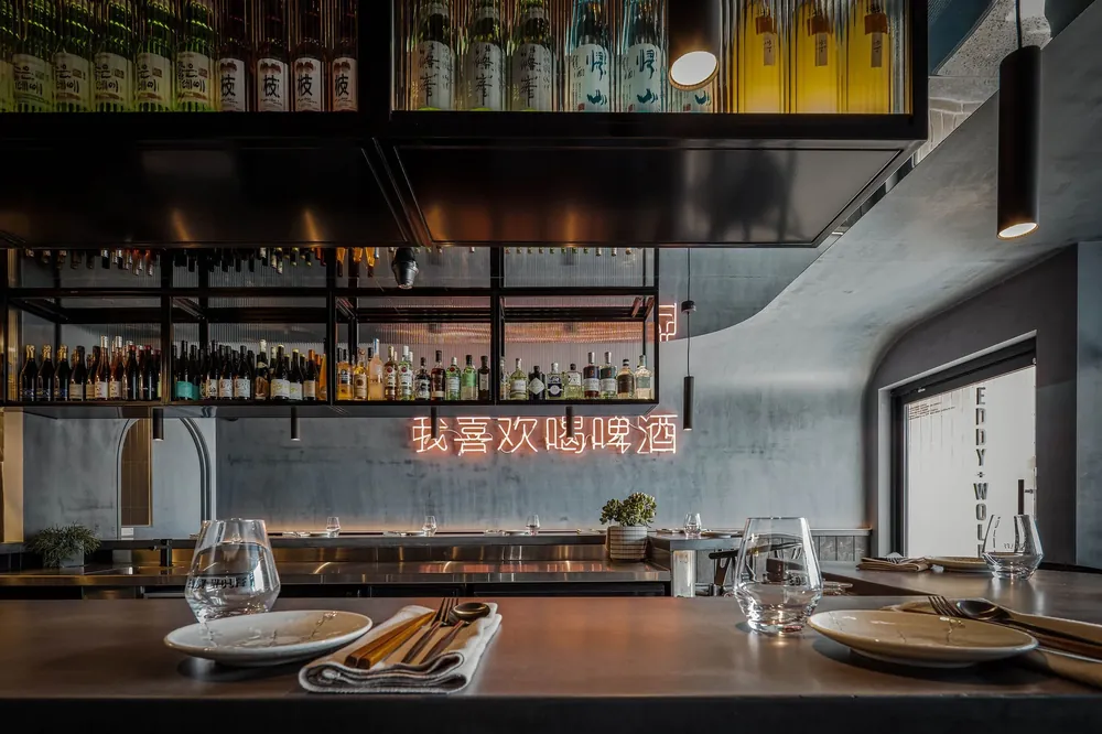 A modern restaurant interior with a bar, tables set with plates, napkins, and glasses, a gray concrete wall, and a neon sign in Chinese characters.
