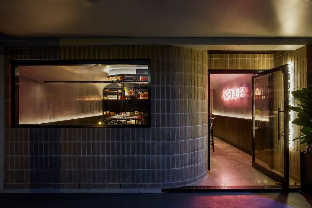 Night view of the entrance to a restaurant with brick exterior, glass window, and open door. Inside, neon signage is visible on the wall, with tables and shelves stocked with bottles and jars.