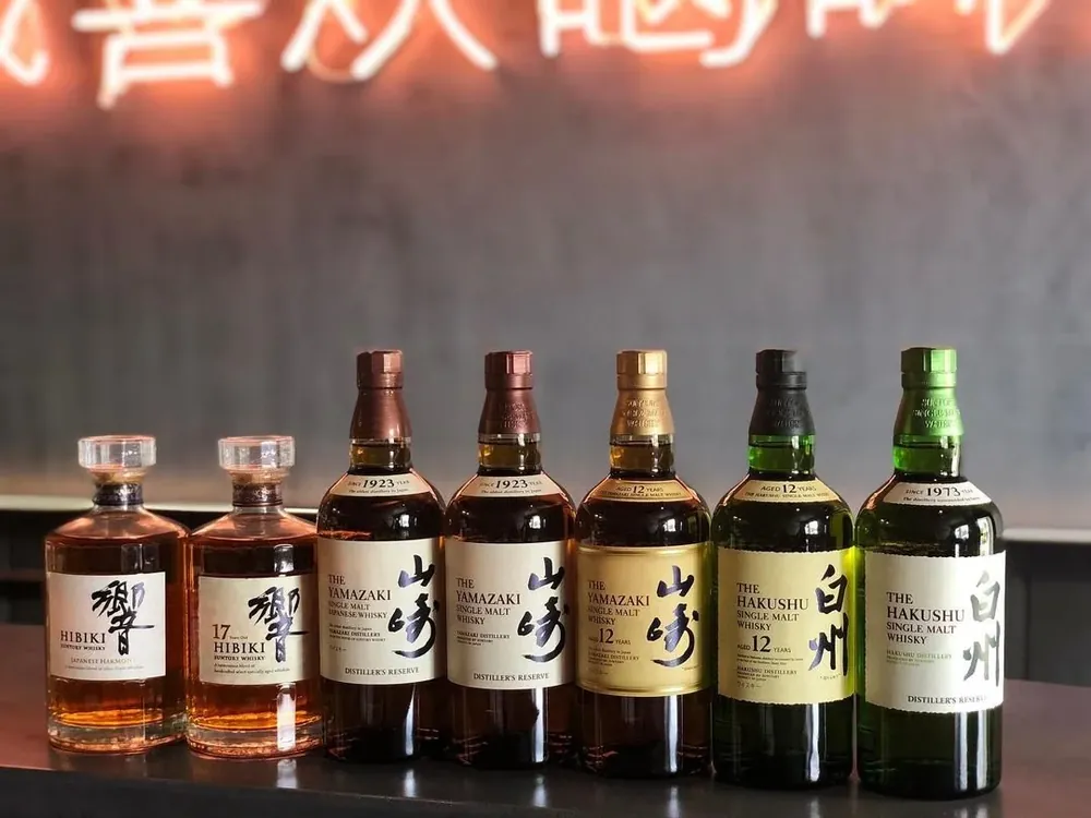 Six bottles of Japanese whiskey, including Hibiki, The Yamazaki, The Hakushu, and their respective labels, arranged on a dark surface with a blurred Asian script sign in the background.