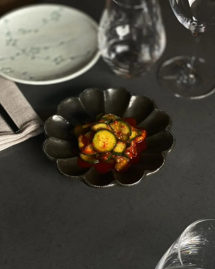 Pickled cucumber with chilli in a dark scalloped bowl, set beside wine glasses on the table